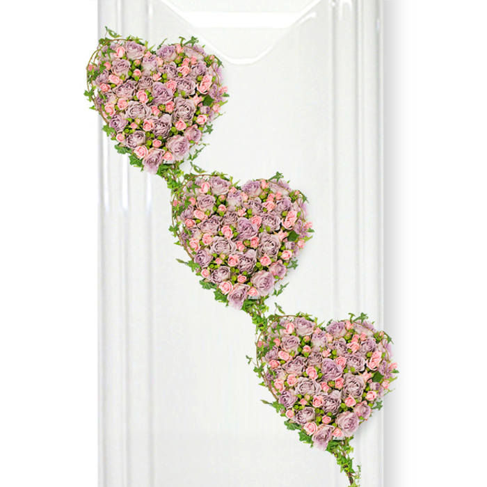In this dignified floral tribute, three perfectly formed heart shapes of roses appear to float one beneath the other, drifting diagonally down across a bright, white setting. Each heart is densely filled with miniature roses, their petals arranged so closely that they create a luxurious, velvety surface. The dominant shade is a soft, dusty lavender-a gentle, almost vintage purple-interwoven with sprays of delicate pastel pink roses that soften and lighten the design. Some blooms are fully unfurled, revealing layered, ruffled centres, while others remain in tighter buds, hinting at the fragrance and freshness of the flowers. Fine green ivy vines trace the outline of every heart, their glossy leaves catching the light as they twist and loop around the floral edges. These vines also link the three hearts together in a subtle vertical cascade, suggesting continuity, shared stories, and a bond that flows from one generation to the next. To the left, faint translucent panels or reflections introduce a hint of glass or polished architecture, evoking the calm interiors of a Canonbury chapel or a quiet Islington venue where families might gather to say goodbye. The overall composition feels serene and composed, with the pastel tones of the roses standing out clearly against the uncluttered background. This design, with its three cascading hearts, would be particularly fitting for expressing love from different members of a family, or marking an enduring partnership, its gentle colours and careful workmanship conveying sympathy, remembrance, and unwavering affection.