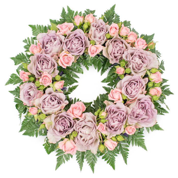 A meticulously crafted floral wreath rests flat on a pristine white surface, its circular form symbolising continuity and eternal remembrance. The arrangement is lush and full, composed primarily of generously petalled roses, each flower carefully angled to create a seamless ring of colour. Dominating the design are large, open roses in a refined blend of dusty lavender and antique mauve, their petals softly shaded from deeper greyish-purple in the centres to paler, weathered lilac at the edges. These cool-toned blooms are interwoven with clusters of smaller blush pink spray roses and tender buds, the pastel pinks bringing a gentle warmth that softens the deeper hues. Together, the roses form a tapestry of texture: ruffled edges, cupped blossoms, and tightly wrapped buds nestling close like shared memories and quiet goodbyes. Beneath and around the flowers, a dense collar of dark green fern fronds creates a structured yet natural base. The ferns, likely leatherleaf or a similar variety, fan out with finely serrated leaves that peek between petals, providing contrast and a sense of depth. Small touches of darker filler foliage are tucked discreetly among the roses, adding dimension without distracting from the main blooms. The wreath is evenly lit, with no harsh shadows, allowing every petal vein and leaf edge to be clearly defined, as if prepared in a calm studio just off Canonbury Road. The overall mood is one of poised serenity and heartfelt respect, suitable for a funeral, crematorium service, or memorial gathering in the Canonbury area, where families seek a floral tribute that honours a loved one with elegance and understated beauty.