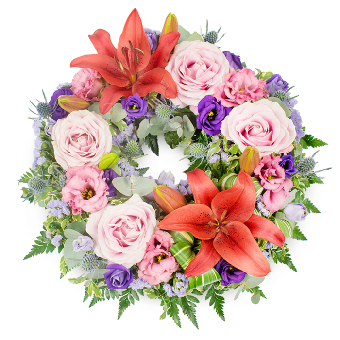 Set against a bright white background, this circular funeral wreath appears richly layered and thoughtfully designed, its flowers closely packed to form a complete ring of colour and texture. Dominating the composition are two large coral-reddish Asiatic lilies, their petals fully unfurled to reveal intricate stamens dusted with pollen. One lily sits proudly near the upper arc of the wreath, while the other anchors the lower portion, creating a gentle visual rhythm that guides the eye around the circle. Between them, clusters of pastel pink and lavender-toned roses are arranged in soft groups, their spiralled petals suggesting grace and heartfelt affection. Ruffled lisianthus, in shades of pale rose, lilac, and rich purple, settle between the roses, providing a cloud-like softness that contrasts with the lilies' bold lines. Fine clusters of lavender-blue filler flowers, similar to statice, are scattered throughout, lending a delicate haze and connecting the different tones. Spiky, blue-grey eryngium thistles are tucked in with care, their sculptural heads bringing subtle drama and a cool, silvery note. The entire wreath rests on a lush bed of foliage: rounded eucalyptus leaves in muted green, feathery fern fronds, and deeper green leaves that frame and support the blooms. A few still-closed lily buds peek out from the greenery, a quiet reminder of life's ongoing cycle. The arrangement feels peaceful yet expressive, the sort of memorial wreath a Canonbury family might order for same-day delivery to a service near Canonbury Station or a farewell gathering around Islington's quiet residential streets, to honour a loved one with beauty and restraint.