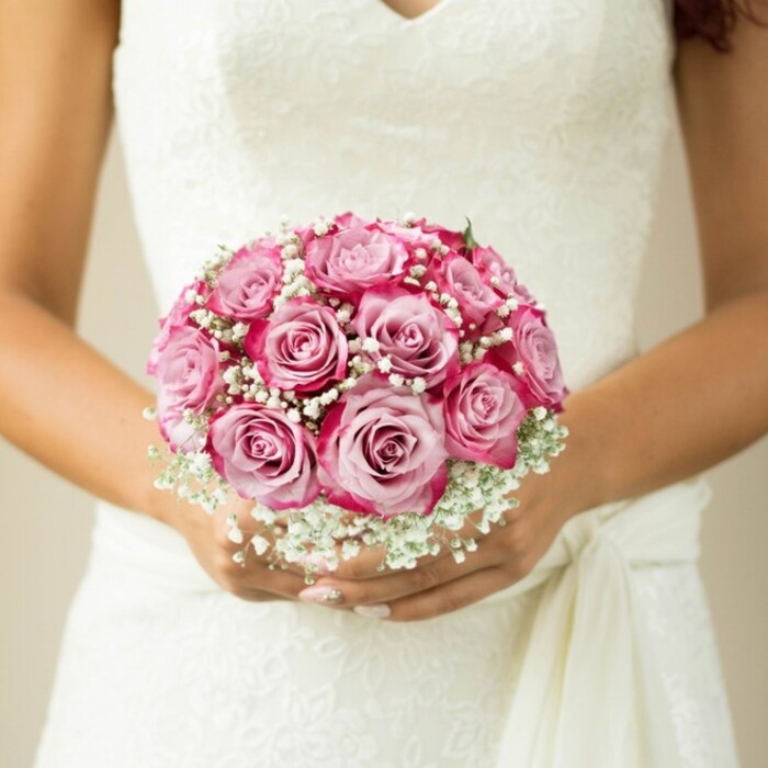 This intimate close-up reveals the hands of a bride amidst the elegance of Canonbury, gently supporting a masterfully arranged bouquet held close to her classic lace wedding dress. With a keen eye for colour, each rose blossoms from a soft, innocent pink at its heart to a jubilant magenta at its margins, the effect undulating in rich contrast beneath the touch of natural daylight. Interspersed among these roses, clouds of white gypsophila spill in airy clusters, imparting both softness and depth, their delicate florets offering a timeless nod to tradition. The bouquet's spherical silhouette speaks to the skill of experienced Canonbury florists, who blend artistry and locality by sourcing the freshest blooms from the area's markets. The bride's elegantly manicured hands-nails refined in a pale, understated hue-cradle the bouquet, while the lace-on-lace bodice and a flowing sash hint at the bespoke craftsmanship underlying the whole ensemble. With the background rendered to a soft, warm haze, every detail of the bouquet's colour palette and textural contrast commands attention. This image resonates with joyful anticipation, making it a perfect reflection of wedding style in Canonbury, whether the celebration takes place along Essex Road or in a hidden leafy garden.