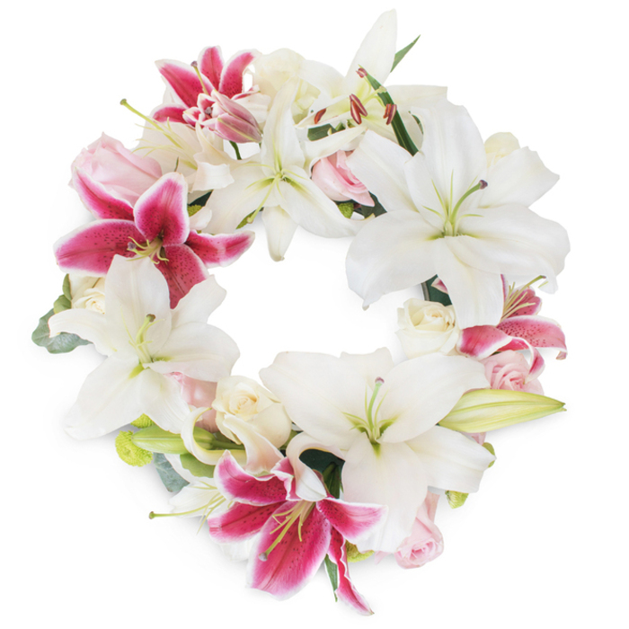 This luminous funeral wreath, lovingly arranged by local florists in Canonbury, epitomises both beauty and reverence in memory's tender hour. A full ring of pristine white Oriental lilies forms the structural heart of the piece, their broad, star-like petals curling gently to reveal pale green stamens and subtle yellow pollen at each open center-symbols of peace, purity, and a life remembered. Interwoven among these, dramatic fuchsia-pink Stargazer lilies impart rich colour, their deep magenta hues tracing vibrant arcs through the arrangement and creating elegant points of focus amid the serenity. Throughout, clusters of creamy white and soft blush pink roses nestle between the larger blooms: some petals are deeply furled in bud, holding stories yet untold, while others have unfurled into full, rounded blossoms suggestive of gentle remembrance. Touches of green come from both glossy lily foliage and wisps of eucalyptus, their muted silver-green shades adding depth, balance, and lightness reminiscent of the leafy paths that wind through Canonbury's gardens. Every flower has been positioned to evoke completeness, the wreath's unbroken circle embodying unity, infinite love, and respectful farewell. Set against a plain white backdrop, the arrangement's pure colours and subtle contrasts glow, each botanical detail elevated into a tribute of artistry and heartfelt condolences. Perfectly suited for funerals, memorials, or the celebration of a cherished life, this arrangement offers support and comfort to Canonbury families, standing as a testament to expert craftsmanship and the enduring spirit of the local community.