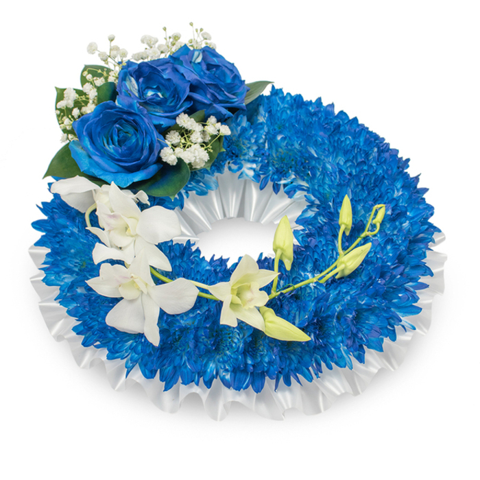 An elegantly composed funeral wreath set against a clean white surface, the main ring densely packed with countless small, vibrant deep-blue chrysanthemum blooms that produce a lush, tactile field of colour. Nestled beneath and framing the blue mass is a ruffled white satin ribbon that traces the inner rim and subtly lifts the flowers, its soft sheen providing a refined contrast to the matt texture of the chrysanthemum heads. A cluster of three large blue roses sits prominently on the upper-left curve; their sumptuous, velvety petals open in a saturated sapphire tone and are accented by tiny white gypsophila sprays and touches of dark green foliage for added depth and a traditional mourner's aesthetic.

Across the lower portion of the wreath a delicate cascade of pristine white orchid flowers drifts from mid-left to lower-right, the orchids' graceful, elongated petals introducing an airy, sculptural quality. Intermingled among these open blooms are pale green orchid buds at differing stages of bloom, lending a quiet sense of renewal within the composition. The interplay of deep blue and pure white conveys solemnity and purity, while the varied textures-spidery chrysanthemums, silky roses, airy baby's breath, and glossy orchids-give the wreath both visual richness and gentle movement. Suitable for commemorative services in Canonbury, near New River Walk or Upper Street, this arrangement expresses respectful remembrance and serene beauty.