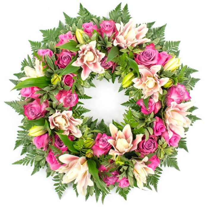 A beautifully composed funeral wreath, captured against a stark white background, forms a perfect circle of colour that immediately draws the eye. The arrangement is lavishly filled with fresh roses in a spectrum of pink tones, from intense fuchsia and magenta to softer blush and pastel shades. Some roses are tightly closed, like small promises of beauty held within, while others are fully open, their many ruffled petals creating a soft, romantic texture around the ring. Intermingled with the roses are large, double-headed lilies, their petals layered and gently curved in tones of creamy white, pale pink, and hinting at peach, adding depth and a sense of movement to the design. Bright lime-green lily buds punctuate the ring at intervals, introducing a lively note that suggests ongoing life and remembrance. Behind and between the blooms, a dense framework of foliage forms a lush green cushion. Fine, feathery fern fronds radiate from the edges, and glossy, broad leaves in rich emerald tones fill any gaps, their smooth surfaces catching the light and contrasting with the softness of the petals. The white background highlights every contour and colour transition, as though the wreath has been set on a workbench in a Canonbury Flowers studio just off Essex Road, ready to be delivered to a nearby service or crematorium. The overall effect is one of tenderness and understated luxury, making this wreath an ideal tribute for funerals, wakes, or memorials in and around Canonbury, where families wish to convey love, gratitude, and a lasting sense of peace through carefully chosen flowers.