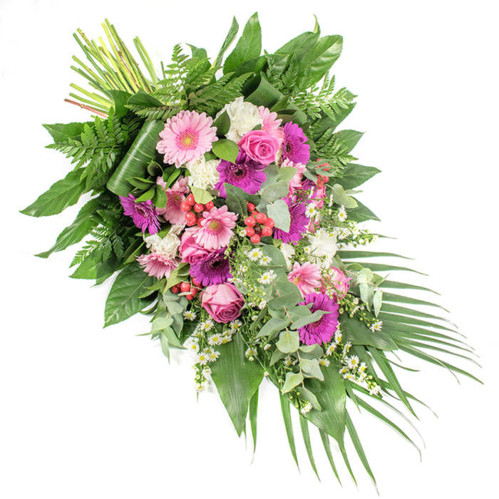 Set against a clean white background, this funeral spray appears as a generous, asymmetrical river of blossoms, moving in a diagonal flow from the top left to the bottom right. It is a richly textured composition that balances vibrant colour with a sense of quiet grace. At its heart, generous gerbera daisies draw the eye-some in pale shell pink, others in deep fuchsia and magenta, each bloom forming a bright, open disc that anchors the design. Interspersed around them are classic pink roses in varying stages of openness, their petals tightly curled at the centre, then gently unfurling outward in carefully layered spirals. Between these larger blooms, clusters of snowy white flowers-likely carnations or small chrysanthemums-add a ruffled softness and help to highlight the surrounding colour. Tiny white daisy-like fillers are threaded throughout, suggesting chamomile or Matricaria, lending a wildflower lightness that feels at home in the leafy streets around Canonbury Square. Pops of glossy red hypericum berries nestle among the petals, introducing a warm accent and a slight textural contrast. The entire spray is supported and framed by an abundance of greenery: wide, dark green leaves provide structure; airy fern fronds create delicate, feathery outlines; and rounded eucalyptus leaves in muted silver-green bring a contemporary touch and hint at a fresh, soothing fragrance. Long, dark palm leaves extend from the lower right, emphasising the cascade and giving the piece a sense of movement. The gathered stems at the upper left are neatly bundled, revealing the care and skill of the florist. Intended as a casket spray or standing tribute for services across Canonbury and nearby Islington chapels, this arrangement speaks of remembrance, respect, and a gentle farewell.