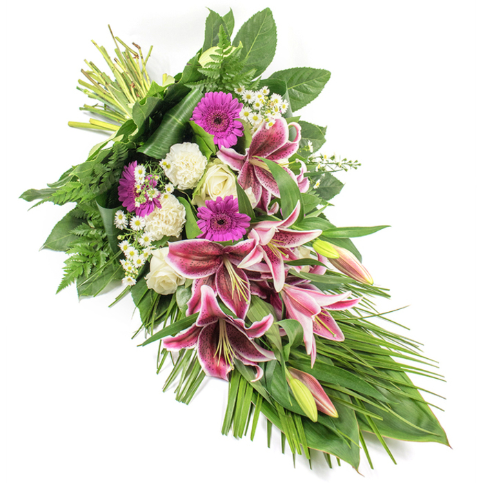 This refined sympathy spray, photographed against a pristine white backdrop, stretches in a graceful diagonal from top left to bottom right, echoing the quiet arc of a farewell. The design is anchored by dramatic Stargazer lilies, their large, star-shaped blooms painted in rich shades of fuchsia-pink, with clean white margins and intricate burgundy speckles that give each petal a velvety depth. A few unopened lily buds, tinted with pale green and soft pink, peek above the open flowers, lending a sense of ongoing life and gentle continuity. Around these, deep magenta gerbera daisies create bold circles of colour, while compact white roses and softly ruffled white carnations bring a sense of innocence and peace. Tiny, daisy-like filler flowers, each petal finely defined, weave between the larger blossoms, adding texture and a cloud-like lightness. The whole arrangement is encircled and extended by an abundance of greenery: broad, glossy leaves provide a solid, dark green backdrop; airy fern fronds whisper at the edges; and slender, grass-like leaves trail elegantly downward, emphasising the elongated, sheaf-like shape. The bound stems at the upper left are tightly wrapped, showcasing skilled craftsmanship reminiscent of a trusted local Canonbury florist preparing tributes for services near Newington Green or quiet ceremonies along Essex Road. The overall impression is of a dignified, heartfelt floral expression, perfectly suited to a funeral service, memorial gathering, or graveside tribute. The blend of strong pinks, tranquil whites, and rich greens evokes both the depth of loss and the enduring warmth of cherished memories.