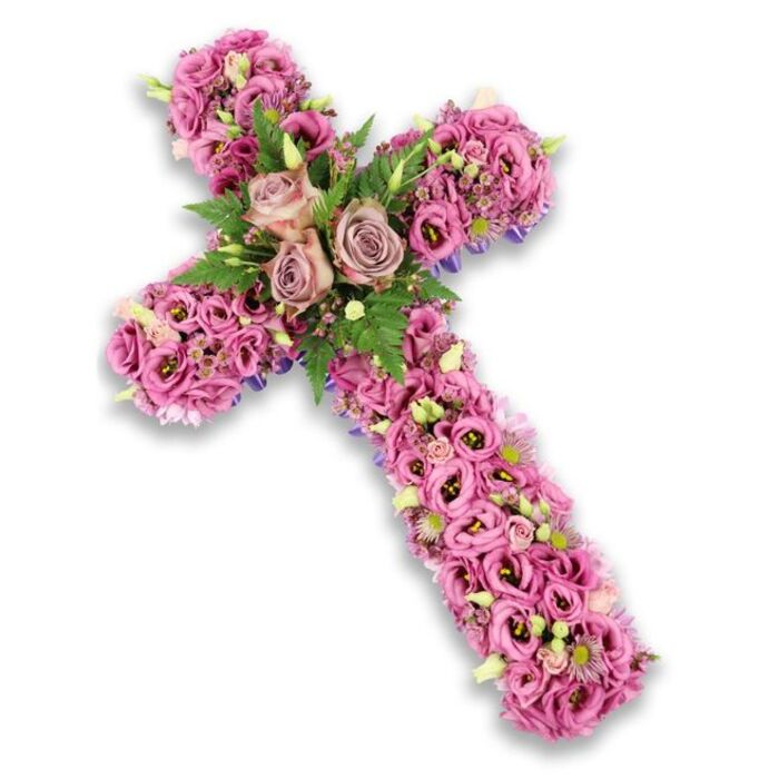 An intricate floral cross arrangement in soft, romantic shades of pink, mauve, and light purple is set against a bright white background, with a gentle drop shadow lending depth to the image. The cross shape itself is completely cloaked in flowers, so generously filled that the surface appears almost quilted with petals. At the central junction of the arms, three statement roses in a muted dusty rose and light mauve tone bloom fully, their spiralled petals forming the emotional core of the piece. They nestle into a collar of textured greenery, including fine fern fronds and small, glossy leaves, whose deep green colour enhances the surrounding pastel palette. Extending outward from this focal point, the arms and length of the cross are lavishly packed with lisianthus blossoms, their trumpet-like forms layered closely together. These lisianthus vary from bright to deeper pink, some with darker centres that add a subtle, shadowed dimension, making the florals feel almost like a living tapestry. Interspersed among them are tiny clusters of pink and lilac fillers, pale green buds not yet opened, and a sprinkling of small white flowers, each one contributing to the intricate texture and rhythm of the design. Along some of the outer edges, slim hints of a darker purple ribbon or base material quietly peek through, helping to define the silhouette without competing with the flowers. The overall effect is peaceful and reverent, the kind of floral cross a family in Canonbury might choose for a funeral, memorial service, or graveside tribute to convey sympathy, remembrance, and a sense of spiritual calm.
