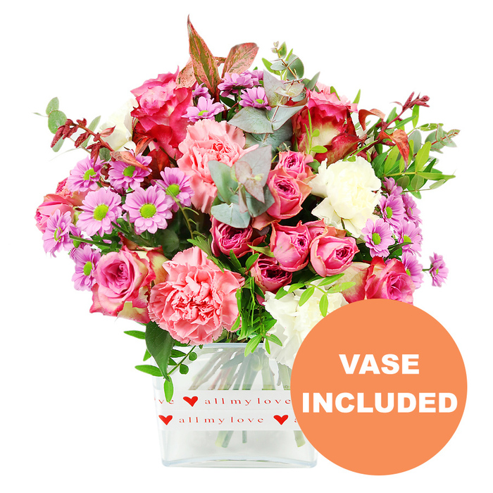In this product photograph, a generous mixed bouquet brimming with pinks, purples, and fresh greens is carefully arranged in a clear, square glass vase and set against a clean, bright white background. The composition is dominated by large pink roses, some in deep fuchsia and magenta tones that darken towards the petal edges, others in soft blush and pastel pink, each bloom layered and velvety, suggesting romance and affection. Interwoven with the roses are full, ruffled carnations in coral, peach-pink, and warm salmon shades, their frilly petals adding fullness and a slightly playful texture. Between the larger flowers, clusters of small daisy-like chrysanthemums in lavender and lilac pop up, each with a vivid lime-green centre that introduces a lively contrast, while a few creamy white blooms help soften and balance the overall palette. The bouquet is wrapped visually by abundant foliage: rounded eucalyptus leaves in a silvery-green hue, deeper green leaves that add depth, and occasional reddish-brown sprigs that offer a subtle hint of autumn warmth. The glass vase itself has gently rounded square corners and a modern, minimalist feel, which is softened by a decorative white band encircling its base. This band is printed with repeated red hearts and the romantic phrase all my love in flowing red script, underscoring the idea of a thoughtful, affectionate gift. In the bottom right corner of the image, an orange circular overlay stands out, showing the words VASE INCLUDED in bold white capital letters to highlight a key feature. The overall effect is of a ready-to-deliver Canonbury bouquet, perfect for sending from a local studio near Canonbury Station to surprise someone in Islington, Highbury, or the surrounding streets with a display that radiates care, celebration, and colour.
