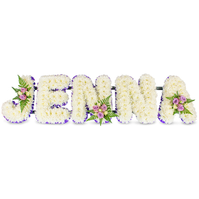 A lovingly crafted floral name tribute reads JENNA in five substantial, three-dimensional capital letters set against a clean white background. Each letter is meticulously built from a dense carpet of small ivory-to-cream chrysanthemums or comparable tiny blooms, producing a plush, cloud-like surface that invites quiet attention. An inner edge of rich purple-either a narrow ribbon or a halo of small lavender florets-maps the negative space within each letter, creating a refined contrast that makes the lettering readable even from a distance. Three artful accent clusters are placed to balance the composition: one on the broad curve of the 'J', another at the lower middle of the first 'N', and a final cluster on the upper right of the 'A'. Those mini bouquets blend soft pink and lavender spray roses with a few pale cream or lemon-tinged roses, threaded through fresh fern fronds that add movement, depth, and a cool green tone. The letters are subtly linked by slender, dark connecting bars which give the tribute a cohesive, structural rhythm suitable for standing display on an easel at a service. Light plays gently across the petals, suggesting an early-morning or late-afternoon glow that lends the piece a contemplative warmth. The scent is quiet and comforting-clean chrysanthemums with a whisper of rose-so the arrangement reads as both a visual and sensory message of affection. Ideal for a funeral or celebration-of-life in Canonbury or nearby De Beauvoir and Newington Green, this personalised floral design communicates reverence, remembrance, and tender admiration.