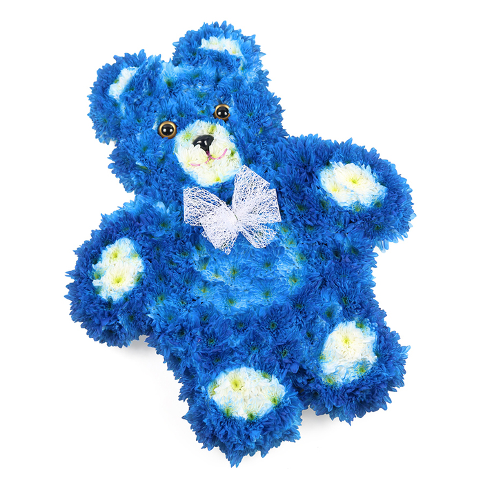 A lovingly crafted floral teddy bear rests against a bright white background, its familiar toy-like outline recreated with meticulous care using live flowers in royal blue and soft cream. From head to toe, the bear is covered in a carpet of small, tightly clustered blue blooms, their petals overlapping to form a dense, cushiony texture reminiscent of plush fur. Subtle variations in the blue shade create depth in the contours of the rounded head, full cheeks, and gently curved arms that seem to open in a silent hug. In contrast, creamy white blossoms define the inner circles of the ears, the rounded snout, and each paw pad, four soft ovals that anchor the design and add brightness to the deep blue body. The bear's face is simple yet expressive: two glossy, dark brown eyes, almost button-like, catch the light, while a small black triangle nose and a fine curved line below it suggest a serene, comforting smile. Around the neck sits a delicate, sheer white bow, its mesh-like fabric looped neatly and slightly ruffled, adding a touch of innocence and ceremony. The whole arrangement appears freshly made, the petals crisp and full, as if prepared that morning in a quiet Canonbury studio for an important farewell. The royal blue hue evokes peace, trust, and remembrance, making this floral bear especially suited to children's funerals, baby boy memorials, or a tender tribute at a crematorium or graveside. For families around Canonbury, Highbury, and the leafy streets nearby, it offers a gentle, symbolic way to express love, comfort, and enduring memory through flowers.