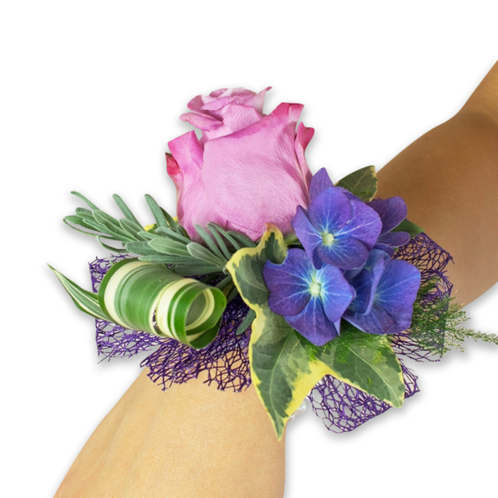 A lively and refined wrist corsage, photographed diagonally on a pale forearm against a stark white background, offers a study in rich contrast and elegant craftsmanship. The central bloom is a generous pink rose, partially open with petals graduating from a concentrated fuchsia at the ruffled edges to a softer, sweet-petal pink at the heart, giving the piece a romantic, luminous focal point. Around it, three dense clusters of deep indigo-purple hydrangea provide sculpted volume; each tiny floret appears to glow from a pale blue-white centre, lending an almost jewel-like depth. Interspersed foliage brings varied textures: long, silvery-green sprigs-reminiscent of eucalyptus or dried lavender-create a soft, airy backdrop while glossy variegated ivy leaves, dark at their core with pale cream margins, add a classical, graceful frame. A modern accent arrives in the form of a broad, white-and-green striped leaf skillfully curled into an architectural spiral, showing the kind of attention to detail we favour at Canonbury Flowers when styling for local events. Below the florals, a dark purple open-weave mesh peeks out, anchoring the palette and introducing a delicate textural shadow. The overall effect is sumptuous yet contemporary, smelling faintly of green stems and cool summer air-ideal for proms, weddings, or celebratory evenings across Canonbury, Essex Road and Upper Street where fresh, handcrafted floral accessories are appreciated.
