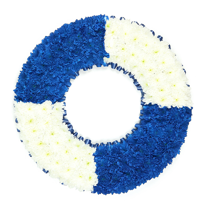 This photograph shows a beautifully balanced floral wreath styled exactly like a lifebuoy, the kind you might associate with riverside walks or coastal towns, yet here reimagined as a tender funeral tribute for families around Canonbury. The arrangement is presented on a stark white background, which throws every petal and curve of the design into sharp relief. The wreath itself forms a perfect circle with a clearly defined hollow centre, every inch densely carpeted with small, button-like blooms that resemble chrysanthemums. The surface appears soft and almost cushiony to the touch, built from layer upon layer of tightly packed flowers. The colour scheme follows a disciplined maritime pattern: from the top left quadrant moving clockwise, the sections alternate between rich royal blue and immaculate white. The blue quadrants are deep and velvety, evoking the inky depths of water beyond nearby London docks, while the white sections feel bright and peaceful, suggesting light breaking over the horizon. Along the inner edge of the ring, a fine border of dark navy ribbon is folded into delicate ruffles, tracing the inner circle like a gentle wave. There is a sense of precision in the spacing and curvature, as though every stem has been measured against the next by a practiced Canonbury florist. The mood is reflective and respectful, suitable for a memorial with links to the navy, lifeguarding, or canal life along the Regent's Canal, but equally fitting as a tribute to someone who offered emotional rescue and support to others. It is a floral emblem of safe passage and steadfast love, ready to be delivered to local services in Canonbury, Barnsbury, or nearby chapels.