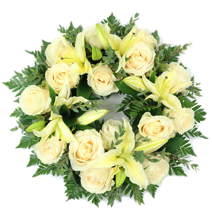 A meticulously composed circular wreath presented flat on a stark white surface, this memorial arrangement marries creamy ivory roses with pale, buttery-yellow lilies against a rich tapestry of green foliage. Around a dozen large, fully bloomed roses - their petals tightly layered and softly luminous - create the primary field of flowers, while several trumpet-shaped lilies in a tender lemon hue are woven through, some opened to display their elongated petals and subtle stamens, others kept in bud to introduce gentle variation in height and texture. The wreath's structure relies on a plentiful navy-to-emerald green base: slender fern fronds give feathery movement around the circumference, and broader, rounded sprigs reminiscent of eucalyptus add cool, muted tones and visual depth that set off the light-coloured blooms. The arrangement is densely packed yet airy, the flowers sitting close enough to read as a single, harmonious surface while still allowing individual blossoms to breathe. Photographed from above in soft daylight, the composition suggests a quiet fragrance - the roses' powdery sweet notes balanced by the lilies' fresher, green-citrus hint - and conveys a mood of solemn grace. Designed for funerals, memorial services, or graveside placement, this wreath is the sort of comforting tribute families in Canonbury and nearby Essex Road often choose when they seek a refined, sympathetic expression of peace, purity, and enduring remembrance.