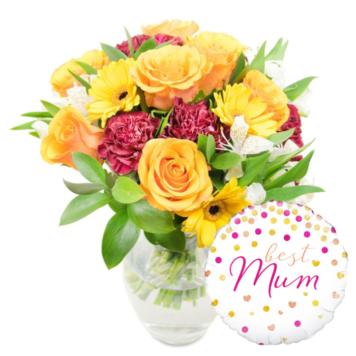A bright and affectionate floral composition, presented against a stark white field, that reads like a carefully chosen gift from a Canonbury neighbourhood shop. The bouquet leans slightly to the centre-left and blooms outward in a tapestry of warm tones: soft orange roses with peach undertones unfurl in rounded, velvety spirals and give the arrangement a refined, classic beauty. Golden-yellow gerbera daisies punctuate the mix, their flat, cheerful faces and dark brown to black centres creating lively contrast. Deep red to burgundy carnations, densely ruffled and plush, add a romantic, tactile density that balances the lighter flowers. Filigree clusters of white alstroemeria - narrow petals with subtle speckles - float among the bolder blooms, offering airy highlights and a gentle scent. A generous cloud of glossy green foliage surrounds and supports the flowers, including broad leaves and slender shoots that add movement and freshness. The bouquet sits in an unfussy clear glass vase so that the viewer can see the trimmed stems and pristine water, a detail that suggests recent, attentive arranging. At the lower right, overlapping the vase and some foliage, a round white foil balloon decorated with gold and fuchsia polka dots and tiny heart shapes proclaims in flowing pink script 'best Mum.' The lighting is soft and natural, the kind that might spill through a window overlooking Upper Street or the Angel on a calm morning, giving the petals a gentle sheen and the whole composition an intimate, celebratory warmth suited to Mother's Day or a special birthday delivery in Canonbury and the surrounding Islington streets.
