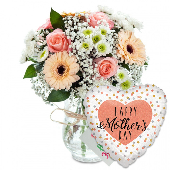 A thoughtfully styled Mother's Day scene is captured against a clean white background, where every shade of peach, pink, and soft green is allowed to sing. To the left, a clear glass vase stands calmly, its jar-like shape wrapped at the neck with a simple strand of light brown twine that gives a hint of rustic charm you might associate with a small Canonbury flower studio. Rising from the vase is a lush, rounded bouquet of fresh spring flowers. Several peach and blush-pink roses sit at the centre, their petals unfolding in soft spirals that suggest both elegance and tenderness. Flanking them are bold peach gerbera daisies, each with a dark brown centre that draws the eye, radiating a bright, optimistic energy. Between these statement blooms, delicate sprays of white gypsophila float like tiny clouds, while small white chrysanthemums with pale green centres add both fullness and a crisp, daisy-like freshness. Slender stems of rich green foliage weave through the design, framing the pastel flowers with natural contrast, much like the greenery lining Canonbury's quiet residential squares. In front of the vase and slightly to the right, a large heart-shaped foil balloon takes up the foreground. Its smooth white surface is dotted with metallic gold and silver spots that shimmer as if catching soft studio light. At the centre of the balloon, a solid peach-coral heart carries the greeting HAPPY Mother's DAY in black lettering, combining bold print with graceful handwritten script. Just behind the balloon, the corner of a small white greeting card peeks out, reinforcing the sense of a complete, ready-to-deliver gift. The overall mood is warm, loving, and celebratory, perfect for a Mother's Day surprise arriving at a Canonbury doorstep.
