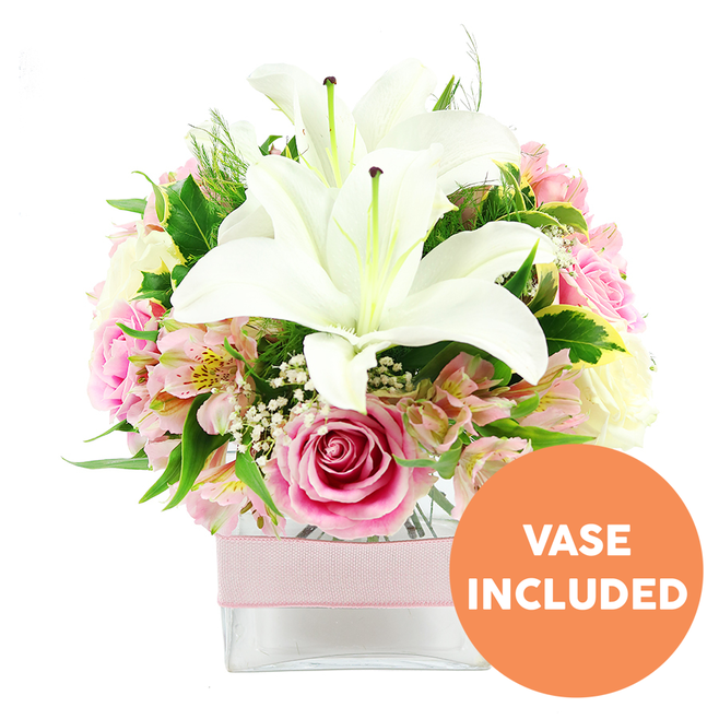 A carefully styled bouquet featuring white lilies and pink roses is set against a spotless white background, ensuring every colour nuance and petal texture can be clearly imagined, as though it were resting on a windowsill in a Canonbury townhouse. At the heart of the display, two generous white Oriental lilies open fully, their petals broad and satin-smooth, curling back to expose bold reddish-brown stamens and the central pistils, creating a striking focal point. Encircling these lilies, an array of pink roses unfolds in layered spirals, some still cupped in tight buds and others fully blossomed into lush, romantic rosettes, conveying the progression of bloom and the passage of gentle time. Between the roses weave graceful stems of pink and white alstroemeria, each floret marked with fine yellow and pink speckles that add painterly detail, while airy clusters of white gypsophila settle into the gaps like soft mist. The palette is grounded by abundant greenery: broad, glossy leaves that catch the light, paired with wispier fronds that introduce texture and movement, echoing the foliage along the quiet paths of the New River Walk. The arrangement is gathered into a low, square clear glass vase, its clean geometry softened by a delicate pale pink ribbon wrapped horizontally around its middle, suggesting a ready-wrapped gift. In the bottom right corner of the image, a round orange badge stands out clearly, bearing the words VASE INCLUDED in bold white capital letters, making the offering unambiguous. Overall, the image presents a fresh, elegant floral piece, ideal for same-day delivery to Canonbury residents to mark a celebration, offer thanks, or brighten a living room near Upper Street with a sense of composed beauty and calm.