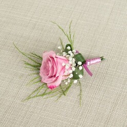 An artfully composed boutonniere rests on a pale, natural linen surface, its woven texture subtly visible beneath the flowers, like a table runner in a town-house near Canonbury Overground station prepared for a wedding morning. At the centre sits a lush blush-pink rose at full, confident bloom, its petals spiralling out from a softly cupped heart. The colour begins as a gentle, powdery pink in the centre and gradually intensifies towards the outer petals into a deeper, more flirtatious magenta, each velvety edge slightly curled as if caught mid-sigh. Encircling the rose's base, tiny white baby's breath flowers cluster in rounded sprays, their numerous pearl-sized blooms forming a cloud-like collar that adds a sense of lightness and romance. Slender blades of fresh green foliage stretch gracefully from behind the rose, some arching outward and some lying closer to the bloom, giving the boutonniere an effortless, slightly whimsical line that would pair beautifully with a modern suit worn to a Canonbury or Highbury Fields ceremony. A broader, darker green leaf is folded with precision around the stems, providing a neatly tailored foundation that mirrors the crisp edge of a lapel. The stems themselves are bound with satin ribbon in tones of lavender and deeper magenta-purple, the sheen of the fabric catching the light in narrow highlights and finishing in a tidy knot rather than a showy bow. This ribbon wrap introduces a subtle accent of colour that harmonises with the rose and gives the design a contemporary edge while staying classically romantic. Soft, natural light falls across the arrangement, creating gentle shadows that emphasise the flower's layers and the fullness of the baby's breath, making this boutonniere feel like a thoughtfully crafted token for weddings, proms, or milestone celebrations across Canonbury and nearby Islington streets.