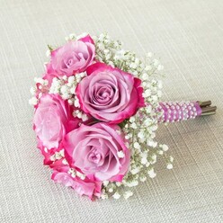 On a pale, almost creamy linen canvas-reminiscent of the sunlit café tables dotted around Canonbury High Street-rests a captivating bouquet that perfectly marries romance with expert craftsmanship. Five radiant roses offer a stunning display of colour gradation, each petal fading from mellow lavender at the centre to an exuberant magenta at the edge, embodying the seasonal beauty found in local gardens near Islington Green. Wisps of white gypsophila, or baby's breath, are threaded between the roses, imparting a sense of airy movement and gentle texture that lifts the bouquet as if floating on a spring breeze. The stems are tightly bundled and sheathed in a luxurious plum-toned satin ribbon, adorned by the spiral of delicate white pearls glinting in the natural light-a detail that hints at tradition but feels wholly modern. The visible ends of the fresh stems add an earthy, organic dimension, underlining the arrangement's authenticity and living vibrancy. Whether destined for a wedding at a Canonbury Road venue or gracing an intimate celebration, this bouquet conjures both innocence and sophistication-a tactile emblem of joy and new beginnings, inspired by the artistry of Canonbury Flowers and their devotion to local floral storytelling.