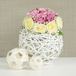 A romantic floral centerpiece is shown in close focus, as if placed on a wedding table in a calm corner of Canonbury, with every petal and texture clearly defined. At the heart of the image is a compact, domed bouquet of roses, roughly a dozen in number, arranged so tightly that the blooms form a smooth, rounded silhouette. The colour palette is soft and harmonious: cool-toned, pastel pink and lavender-rose heads mingle with buttery cream and pale yellow roses, their petals gently spiralling outward. Each flower looks freshly cut, like something a careful florist would prepare early in the morning before delivering to a celebration near New River Walk. Between the roses, airy stems of white baby's breath weave through the arrangement, creating tiny constellations of blossoms that introduce lightness and movement. Supporting this bouquet is a distinctive spherical vase, crafted from slim white wicker or branch-like strands, crossed and knotted into an open, cage-like globe. Through the gaps of this white lattice you can just glimpse the inner container, adding depth and a handcrafted charm that feels both modern and rustic. The vase stands on a smooth white surface, uncluttered and bright, which helps the subtle colours of the flowers gently glow. To the left sit two decorative orbs made from pale, natural wood or rattan; their open weave and differing sizes subtly mirror the main vase and complete the tableau. Behind, a softly textured beige background, reminiscent of fabric or finely woven wallpaper, keeps the overall look warm and understated. The composition conveys quiet luxury and intimate celebration, ideal for illustrating elegant wedding table flowers, anniversary centrepieces, or refined home décor ideas for Canonbury homes.