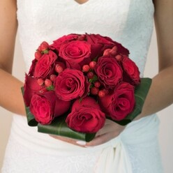 A sense of classic North London romance breathes from this detailed portrait of a bridal bouquet, imagined and styled by the artisans at Canonbury Flowers. Bathed in soft, natural light - perhaps reminiscent of an early afternoon by the New River Walk - a bride's hands cradle a bouquet imbued with drama and elegance. The stately, domed arrangement bursts with velvety, deep red roses, their petals overlapping in gentle whorls from the tight embrace of new buds to blossoms confidently unfurled. Throughout the roses, bright red hypericum berries glimmer like precious rubies, adding tempting texture and a splash of festive intrigue to the bouquet's tapestry. Surrounding the blooms, sculpted broad green leaves are expertly wrapped, their glossy finish outlining the flowers and grounding the arrangement in a lush, organic frame. Creamy lace and fine details of the bride's white wedding dress can be glimpsed, hinting at an atmosphere of tradition and refinement against the softly blurred background. The bouquet's fullness seems to promise unity and abundance, its fragrance conjuring up whispers of vows exchanged in Canonbury's historic chapels. With every detail - from manicured fingertips to the careful play of color - this bouquet becomes not just a floral centerpiece, but a cherished memory for any local celebration or bridal editorial reflecting the artistry and heritage of North London.