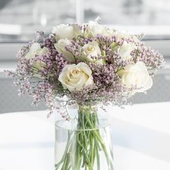 This detailed image shows an elegant sympathy arrangement of fresh white roses and ethereal purple filler flowers, composed with the kind of care a Canonbury florist would bring to a sensitive occasion. At the centre of the frame, generous ivory and creamy white roses unfurl into open rosettes, their petals layered in soft spirals that evoke both innocence and quiet strength. Nestled between the larger blooms are fine sprays of lavender and lilac statice, their tiny, cloud-like florets lending a misty, almost dreamlike texture that gently breaks up the smoothness of the rose petals. Here and there, the filler flowers carry a blush of pale pink, which adds a touch of warmth while preserving the overall cool, tranquil palette. The arrangement sits in a tall cylindrical glass vase, crystal clear so that every green stem is visible as it descends into the clean water; this glimpse of structure beneath the surface hints at the florist's skill and the freshness of the flowers just prepared for delivery. The bouquet rests on a spotless white tabletop, similar to a sideboard in a calm Canonbury living room or a quiet study off Essex Road, catching soft natural light that skims across the blooms and picks up subtle highlights on the petals and the glass. The background is gently out of focus in pale neutral tones, giving the impression of an airy, contemporary interior. Overall, the composition conveys solace, respect, and a peaceful grace, making it an ideal expression of sympathy for families in Canonbury, Highbury, or nearby Islington who wish to send a dignified, comforting floral tribute.