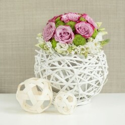 A round white woven vase, shaped like a gently flattened sphere, sits centered on a crisp white tabletop, its open rattan-style strands creating intriguing shadows and a feeling of lightness. Rising from this sculptural base is a lush, domed bouquet of pastel blooms, as carefully composed as a table setting for an intimate Canonbury wedding reception. Soft pink and lavender roses, many fully unfurled, command attention; some petals deepen into a more vivid rose pink at the edges, giving each flower a unique personality. Between these, vivid green button chrysanthemums peek out, their tight, textured heads offering a playful contrast to the roses' smooth, spiralling petals and adding a fresh, garden-like energy. Around the lower edge of the dome, small white filler flowers weave in and out, slightly draping over the lip of the vase as if gently spilling towards the table, lending movement and a relaxed, natural grace. To the left, two decorative wicker-like spheres, one larger and one smaller, rest on the white surface, their pale, interlaced strands echoing the vase and reinforcing a theme of craftsmanship and organic design familiar to local Canonbury florists. The softly blurred beige background, reminiscent of woven fabric or a lightly textured wall, keeps the focus firmly on the flowers while bathing the scene in warmth. This arrangement feels ideal as a refined table centerpiece for a registry office celebration near Highbury & Islington, or a stylish dinner in a Canonbury Garden apartment, radiating gentle romance, freshness, and understated sophistication.