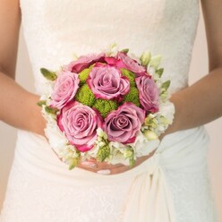 This evocative bridal moment unfolds in the heart of Canonbury-a serene close-up of a bride holding a luxuriously rounded wedding bouquet, perfectly paired with her vintage-inspired gown. The dress, likely in an off-white or creamy ivory tone, features ornate lacework that caresses her arms and bodice, the details illuminated in soft, natural light reminiscent of a quiet walk along Canonbury Road. Central to the ensemble, plush roses in gentle dusty mauve and lavender hues form the bouquet's focal point, each bloom edged with deeper notes of fuchsia that create a painterly play of colour. Amidst these romantic blooms, vibrant lime-green pom-pom chrysanthemums add a burst of cheerful energy, their compact, textured spheres reminiscent of the lush green spaces of Highbury Fields nearby. This radiant core is framed by a halo of small, creamy-white blossoms-perhaps sweet-scented freesias or petite spray roses-whose airy presence bestows the bouquet with an unmistakable sense of bridal grace. Fresh green buds and foliage lend movement and depth, peeking out between blooms for a look both natural and curated. The bride's carefully pampered hands, nails painted a gentle blush with a touch of subtle sparkle, cradle the bouquet with tenderness and poise. Bathed in gentle midday light, every detail comes alive-the texture of lace, the dewy freshness of petals-all telling the story of a Canonbury celebration: intimate, joyful, and styled with a true florist's expertise, ideal for weddings in Islington and surrounding neighbourhoods.