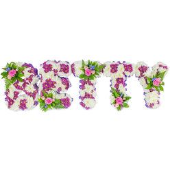 A quiet, composed floral tribute spells out BETTY in strong, block letters, each letter a tactile study of white and purple against a pristine white backdrop. The main bodies are formed from close-set creamy white chrysanthemums that create a plush, cushiony surface-soft to the eye, like fresh snow under morning light. Within those white masses a secondary pattern of deep fuchsia to magenta-purple chrysanthemums is used to outline and sculpt the letters, producing a refined contrast so the name reads from a distance yet invites a closer gaze. Along the sides a slim purple ribbon traces the silhouette, lending a ribboned seam that echoes the purple blooms. Carefully positioned accent clusters punctuate the design: small groupings of gentle pink roses, from tight buds to emerging blooms, sit atop the B, the center T, and the upper arm of the Y. These roses are paired with tiny sky-blue fillers-delphinium or cornflower-like sprigs-and soft, lacy ferns that bring an airy green texture into the arrangement. The overall palette-white, purple, pink and blue-feels both respectful and tender, meant to comfort rather than overwhelm. I imagine this bespoke tribute placed on a polished stand at a Canonbury funeral service near Upper Street, the floral name offering a clear, loving marker of memory. As an experienced local florist I note that this composition balances traditional sympathy colours with contemporary detailing, a piece that speaks softly when words are scarce and honours a life with careful, handcrafted restraint.