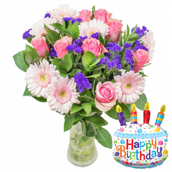 Bathed in soft, neutral light against a crisp white backdrop, this birthday arrangement blends classic romance with a touch of party-ready fun. The dominant element is a lush cluster of soft pink roses: petals layered like silk, ranging from pale blush at the edges to richer pinks toward the center, arranged to create a rounded, generous silhouette. Complementing the roses are pale pink Gerbera daisies whose broad petals and pale yellow centers open like friendly faces, giving the bouquet cheer and presence. Scattered throughout are concentrated sprays of vivid purple statice (limonium), their textured, papery blooms creating pockets of contrast and a painterly sense of depth. Small white filler blooms and abundant glossy green foliage support the composition, framing each flower and lending a natural, garden-fresh feel. The stems rest in a transparent glass vase with a slightly wider base so water and green stalks are visible, emphasizing freshness and careful handling. Nestled to the right, a cake-shaped foil balloon adds celebration: white frosting with wavy blue piping, colourful sprinkles, and four multicoloured candles-depicted purple, red, orange and blue with yellow flames-above a playful "Happy Birthday" message spelled in bright letters; a tiny "KALEIDOSCOPE" mark is visible at the bottom. As a Canonbury florist, I imagine sending this as a birthday surprise for neighbours near Canonbury Square or deliveries across Essex Road and Newington Green-an arrangement that reads as intimate, jubilant, and freshly prepared by Canonbury Flowers for a loved one's special day.