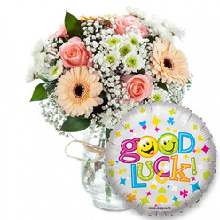 A sunlit, supportive gift set sits against pure white: a lush bouquet arranged slightly left-of-centre in a simple clear glass vase, its neck finished with rustic twine that speaks of handcrafted care. The flowers combine soft peach and pale pink roses with the round, optimistic faces of peachy-orange Gerbera daisies, each with a contrasting dark brown eye that anchors the palette. Cloud-like swathes of white baby's breath (Gypsophila) weave through the stems like airy lace, while tight clusters of small white and lime-green chrysanthemum-type florets introduce a crisp, lively texture. Glossy green foliage rises between blooms, lending shadow and a subtle leafy perfume to the bouquet. Prominently placed in the right foreground and overlapping the lower vase, a reflective silver Mylar balloon catches the light; its metallic ground is dotted with colourful stars, hearts and tiny geometric shapes in blues, pinks, yellows and oranges. Across the balloon the phrase "Good Luck!" appears in large, playful multi-coloured letters - the two 'o's in "Good" replaced by bright yellow smiley faces (one winking, one playfully sticking out its tongue) that give the gift an affectionate, human touch. This image, styled for clarity and warmth, evokes sending encouragement across short distances in Canonbury - whether to a student heading to an exam near Newington Green, a friend starting a new job on Essex Road, or a neighbour taking a brave step - a composition we often assemble at Canonbury Flowers for local delivery.