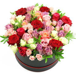 A richly romantic rose hatbox arrangement fills the frame, captured from above and slightly to the side so the viewer can appreciate both the rounded shape of the bouquet and the smart container that holds it. The flowers surge upwards in a full dome, overflowing a sleek black cylindrical hat box edged with a narrow ribbon of red at the base, giving a nod to classic luxury gift packaging reminiscent of chic boutiques off Upper Street in Canonbury. The mix of roses is striking: large, velvety deep red blooms sit prominently, their petals layered and plush, radiating passion and drama. Threaded through them are fuchsia and soft blush pink roses, peach and coral tones that bring warmth, and elegant cream and ivory flowers that soften the palette. Hints of lavender and pale purple roses run through the arrangement, lending a slightly contemporary, almost Parisian feel. Some heads are fully open, showing every curve of the petals, while others remain partially closed or appear as smaller spray clusters, creating texture and depth. Sprigs of fresh green foliage, perhaps ruscus or eucalyptus, peek between the blooms, the cool green leaves keeping the design from feeling too heavy. Set against a pure white background and lit with bright, even light, every shade and petal edge is clearly defined. The result is a bouquet that feels both generous and meticulously composed, the kind of premium rose hatbox Canonbury Flowers might send for a special anniversary dinner near New River Walk, a milestone birthday, or a heartfelt thank-you delivered the same day across N1.