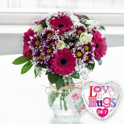 An elegant yet playfully romantic floral display is captured in bright, natural light, as if placed on a windowsill in a Canonbury townhouse overlooking a quiet tree-lined street. At the heart of the image is a rounded bouquet dominated by generous gerbera daisies in intense magenta and fuchsia, their bold daisy heads radiating around dark central discs that draw the eye. Weaving between these statement blooms are smaller, multi-toned chrysanthemums whose petals shift from deep purple near the centre to crisp white at the tips, each flower punctuated by a golden yellow core that adds a cheerful sparkle. Creamy white roses nestle in among the brighter colours, their layered petals smooth and almost velvety, bringing a touch of classic elegance. Airy clusters of white baby's breath soften the transitions between each blossom, lending a feathery lightness that keeps the bouquet from feeling heavy. Lush green foliage adds depth and contrast, framing the flowers and hinting at a fresh, garden-like scent. The arrangement stands in a clear, rounded glass vase, the tidily cut stems and clear water visible beneath, giving the composition a clean, modern look. Around the neck of the vase, a band of natural twine is tied, supporting a small white tag decorated with a simple pink rose motif and friendly green writing that suggests the bouquet has been lovingly gathered by a local florist. To the right, completing the gift, a gleaming silver-edged, heart-shaped foil balloon with a white centre showcases colourful, patterned letters spelling "Love n' Hugs 24-7," echoing the affectionate theme. Set against a pale surface and a softly blurred, luminous background, the scene feels like a thoughtful delivery arriving to a home near Canonbury Station, perfect for birthdays, anniversaries, or sending a warm hug in floral form.