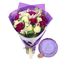 A lush romantic bouquet with a matching balloon is shown against a bright white backdrop, as if waiting on a counter in a Canonbury florist just off Essex Road. The flowers are gathered in a generous hand-tied cluster, led by soft cream-white roses whose petals curl inward in tight spirals, suggesting freshness and the calm anticipation of a special evening. Woven among them, velvety carnations in deep magenta and burgundy tones create pools of colour, their ruffled, feathery edges adding volume and depth. Slender stems of white alstroemeria slip between the larger blooms, their narrow petals marked with fine reddish-brown streaks and freckles that bring a subtle, intricate patterning to the design. Around the flowers, lush greenery in varying shades of green, including some leaves edged with pale cream, shapes the bouquet like a natural frame. The florist has wrapped the stems in two layers: an inner band of crisp white paper for clean structure and a sheer purple cellophane outer layer that fans up around the blooms, crinkled and semi-transparent, catching the light in gentle, lilac reflections. A slim, bright green satin ribbon holds the base, tied in a neat bow with a simple, rectangular brown tag dangling from it, ready to be inscribed with a message from a local Canonbury partner. To the right of the bouquet lies a small, metallic purple foil balloon, round with a scalloped, rosette-style edge. Its centre circle is a lighter lavender shade, where the scripted words "To Loving Partners" appear in a soft pink or magenta hue. A tiny rainbow holographic sticker hugs one side, hinting at an iridescent sheen. The whole ensemble feels tender and celebratory, perfectly suited to anniversaries, date nights at home, or to mark shared milestones for couples living around Highbury & Islington and the leafy squares of Canonbury.
