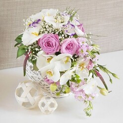 An artfully composed wedding-style centerpiece is captured in a luminous close-up, featuring a round white wicker basket overflowing with romantic pink and white flowers, as if prepared by an experienced Canonbury florist for an intimate reception on a leafy side street off Upper Street. At the heart of the arrangement, large pastel pink roses unfurl in perfect spirals, their layered petals giving a plush, velvety texture that instantly suggests tenderness and celebration. Around them, wide-winged white Phalaenopsis orchids hover like butterflies, each bloom showcasing a warm yellow and subtle orange centre, which adds depth and elegance to the overall palette. Between these focal flowers, ruffled white lisianthus, some with a delicate purple-tinted edge, introduce gentle movement and a slightly wilder, garden-grown charm. Pale pink stock flowers extend from the basket's edge, their clustered florets creating soft vertical lines that seem to gently cascade, echoing the flow of a bridal gown. Fine sprays of white baby's breath are dotted throughout, giving a misty, almost ethereal quality, as though the blooms are suspended in air. Fresh green foliage threads through the design, with slim leaves and tender buds breaking up the softer tones and adding a freshly picked feeling, like flowers gathered from a hidden Canonbury garden. Resting beside the basket on a bright white surface are two light wooden or rattan spheres, their open-weave patterns mirroring the basket's craftsmanship and reinforcing the rustic-yet-refined mood. Behind everything, a soft, neutral fabric backdrop in warm beige allows the arrangement's colours and textures to stand out clearly, evoking a sense of calm elegance and making this scene ideal for illustrating wedding table arrangements, anniversary dinners, or romantic home décor in and around Canonbury.
