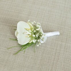A refined white orchid boutonniere lies gently on a pale beige, woven fabric surface, the texture reminiscent of natural linen one might find in a stylish Canonbury townhouse near Essex Road. The central focus is a single Phalaenopsis orchid, its petals a flawless, luminous white that curves outward in a soft, sculptural shape. The inner throat of the orchid glows with a delicate wash of pale yellow and creamy ivory, adding a hint of warmth, like late afternoon light filtering through London townhouse windows. Surrounding the orchid is a delicate collar of baby's breath; countless tiny Gypsophila blossoms form a misty, cloud-like ring, their minuscule petals clustered on fine stems that lend an ethereal lightness to the buttonhole. Interspersed among them are strands of fine green foliage, perhaps maidenhair or asparagus fern, whose feathery fronds arc outward and give the design a gentle sense of movement and freshness, as though just gathered from a shaded garden off Canonbury Square. The stems of the orchid, Gypsophila, and greenery are tidily bound in a band of crisp white satin ribbon, wrapped with even tension so it lies smooth and flat, finished with a discreet, elegant bow at the base. The soft, diffused lighting in the image enhances every subtle texture: the satin's understated sheen, the orchid's velvety surface, and the fabric's faint weave. The result is a boutonniere that feels quietly luxurious and deeply romantic, ideal for a modern London wedding, a civil ceremony at a nearby Islington venue, or any formal event where a touch of pure, white floral elegance is desired on the lapel.