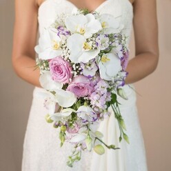 Envision a serene bridal moment unfolding on a peaceful street near Canonbury Square, where a bride, shown from the mid-waist upwards, cradles a breathtaking bridal bouquet crafted in a classic cascading style. The bouquet commands attention with its sculptural teardrop silhouette, dominated by ivory phalaenopsis orchids whose dewy petals softly reflect the neutral, sunlit backdrop. Cupped within these orchids are lush pink garden roses, each bloom unfurling in velvety layers that echo the gentle blush cast of early morning light over Canonbury's townhouses. The floral composition is thoughtfully interlaced with slender lavender stock flowers, their spiky forms introducing whimsical movement, while ruffled white lisianthus-some edges tinted in deep amethyst-provide intricate contrast. Threads of airy white gypsophila add a sense of dreamy volume, while minimal leafy sprigs impart an organic finish without overpowering the soft palette. The bride's white gown, adorned in whisper-fine lace along the bodice, subtly frames the flowers, her hands gently supportive beneath the bouquet's natural waterfall. It's a bouquet that blends texture and fragrance, conjuring the feeling of a fragrant morning stroll past Canonbury's charming gardens. Evoking both gentle anticipation and timeless romance, this bridal arrangement is a testament to expert floristry and Canonbury's unique blend of heritage and elegance-perfect for the most graceful wedding ceremonies in this beloved neighbourhood.