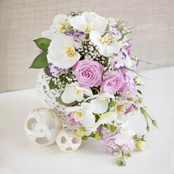 A refined floral centerpiece, ideal for an elegant Canonbury wedding table, sits serenely on a smooth white surface, bathed in soft, even light that reveals every nuance of colour and texture. Dominating the design are several large white Phalaenopsis orchids, their petals broad, gently curved, and almost porcelain-like, each bloom centered with a touch of golden yellow and soft amber that creates a subtle glow amid the cooler shades. Around them, pastel pink and lilac roses are gathered in full, velvety rosettes, some fully open and some still cupped, suggesting a sense of unfolding romance that would suit a reception near Canonbury Gardens or a chic celebration off Essex Road. Interwoven with the roses, frilled Lisianthus in white and purple tones add depth, their lightly ruffled petals giving a sense of movement, while clouds of delicate white baby's breath drift between the larger flowers, softening every edge and filling the bouquet with a touch of dreamlike lightness. Slender stems bearing pale purple blooms cascade gently downward, lending a natural, flowing line that draws the eye from the top of the design to the table below. The flowers are gathered into a striking white spherical vase with an open, lattice-like weave, reminiscent of fine wicker or rattan, which gives the whole composition a contemporary, textural character. Beside the main arrangement sit two cream-toned decorative spheres, similarly woven, one slightly larger than the other, echoing the vase's pattern and reinforcing the sense of balance and thoughtful design. The neutral, softly textured background melts away behind the display, making the fresh colours and lush greenery feel especially vivid-an artful floral statement worthy of a Canonbury townhouse dinner or an intimate engagement party.