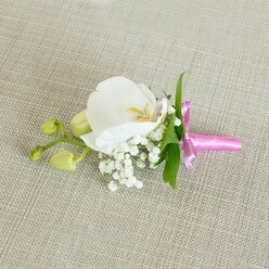 A refined floral boutonniere is shown resting lengthways on a pale beige, linen-textured fabric, its understated background reminiscent of the calm, light interiors of Canonbury townhouses. At the heart of the design sits a single white Phalaenopsis orchid, fully open and perfectly formed. Its broad, silky petals unfold gracefully, their pure white surface catching the soft, even light. In the centre, the orchid's throat reveals subtle shading - a gentle wash of soft yellow and the faintest whisper of blush pink - adding depth and a hint of warmth without disturbing the bloom's clean simplicity. Framing the base of the orchid is a delicate ring of baby's breath, each tiny white flower forming a soft, airy cloud that lends an almost dreamlike quality to the piece. Fresh, slender green leaves peek out from behind, their smooth, slightly glossy surfaces offering a natural counterpoint to the more delicate textures of petals and gypsophila. To one side, a short stem extends outward, bearing a pair of tightly closed lime-green orchid buds that speak of new beginnings and the promise of future flowering - a tender symbol for a wedding, civil ceremony, or milestone celebration in and around Canonbury, perhaps near Islington Green or alongside a registry office gathering. The stems are neatly bound in a vivid fuchsia pink satin ribbon, carefully wrapped and tied into a small, precise bow. The ribbon's bright colour and light-catching sheen introduce a joyful, contemporary flourish, standing out beautifully against both the neutral linen and the white florals. Illumination is gentle and even, accentuating the smooth planes of the orchid, the tiny spheres of the baby's breath, and the weave of the fabric. It feels like a piece prepared with quiet concentration by an experienced local florist just before it is pinned to a lapel for an evening of celebration.