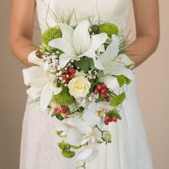 Against the serene setting of a Canonbury wedding, a bride tenderly holds a sumptuous, cascading bouquet, its artistry echoing the refined charm of Islington's leafy lanes. The design is a masterful blend of textures and tones: bold white lilies, each petal dusted faintly with reddish-brown flecks, offer a star-like focal point reminiscent of honeysuckle fragranced evenings in local gardens. Nestled at the heart, a classic creamy rose unfurls with subtle sweetness, setting a gentle, romantic tone. The bouquet's signature teardrop silhouette is shaped by a graceful trail of white phalaenopsis orchids, their petals catching soft daylight to impart effortless elegance and movement. Scattered throughout, lime-green cushion mums mirror the vibrancy of New River Walk's spring foliage, lending a cheerful, modern twist to the palette. Plump, ruby-red hypericum berries glimmer amidst a delicate haze of baby's breath, creating bursts of festive colour and joyful contrast seen in Canonbury market bouquets. Fine sprays of airy greenery and longer wisps encircle the blooms, offering an organic, free-flowing frame. The bridal gown's delicate lace-just visible where her hands gently embrace the arrangement-adds layers of romance and intimacy. The background, softly out-of-focus, centers attention on the intricate bouquet, making it an inspiring choice for Canonbury ceremonies or features on modern London weddings.