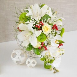 An opulent white and green floral centerpiece is displayed on a pristine white tabletop, set against a softly textured beige backdrop that feels like the calm, neutral walls of a Canonbury dining room. The arrangement rises from a round white woven basket, its wicker strands forming a subtle pattern that complements the organic lines of the flowers. Several large white lilies dominate the upper half, their trumpet blooms open wide, petals gently curling back to reveal delicate speckles and pale golden stamens that suggest a light, sweet scent drifting across the table. Nestled among them is a single cream-yellow rose, perfectly formed and slightly cupped, creating a warm focal point that draws the gaze inward. From the right side, a graceful cascade of white phalaenopsis orchids spills downward, each blossom with rounded petals and a soft, sculptural shape, forming a bridal waterfall that would suit a wedding breakfast near Canonbury Square. A lush base of greenery holds everything together: fine, feathery leaves, slender grasses, and broader glossy foliage that introduce layers of texture and depth. Bright green pom-pom blooms, reminiscent of mini green dianthus, pop up between the stems like little cushions, contrasting beautifully with the smooth orchid petals. Threads of white baby's breath wind through the arrangement, while clusters of rich red berries add festive sparks of colour and a slightly woodland feel, as though freshly gathered from the edges of Highbury Fields. To the left of the basket, two delicate white decorative spheres, made from intersecting strips that form open lattice balls, echo the basket's weave and add a sculptural touch. The entire design radiates quiet celebration, ideal for a wedding table, anniversary dinner, or elegant gathering in Canonbury, combining purity, romance, and refined craftsmanship in one thoughtful composition.