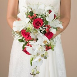 A bride, partially out of focus yet radiantly poised, cradles a grand, teardrop-shaped bouquet in front of her softly illuminated white lace gown, her bare arms embracing the ensemble with calm anticipation. The bouquet, envisioned by a skilled Canonbury florist, showcases open white lilies at the center, their broad petals dramatically fanning out and golden stamens glinting in the ambient light, filling the air with a graceful sweetness. Around them, trailing stems of elegant phalaenopsis orchids gently descend, adding flowing motion and a hint of modern romance inspired by wedding scenes in the tranquil gardens between Astey's Row and Canonbury Square. Rich red roses, their lush, velvety petals deeply saturated, intertwine with sprays of soft baby's breath and a scattering of ruby hypericum berries, casting bright, festive accents. Wisps of fine asparagus fern and gleaming foliage weave throughout the design, grounding the bouquet in nature. The composition is both airy and structural, a harmonious blend of texture and hue that mirrors the quiet sophistication of the Canonbury community. Hints of spiky greenery peek through, lending depth and a sense of the unexpected. The neutral background recedes into gentle softness, ensuring that the bouquet's vibrant colors and the bride's gentle posture remain the heart of the image. As the memorable centerpiece of a Canonbury wedding, this bouquet embodies timeless beauty, joyful beginnings, and the attentive touch of local artisan floristry.