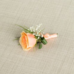 A single peach rose boutonniere is beautifully showcased on a pale beige linen-style fabric, its fine, even weave forming a gentle, neutral backdrop reminiscent of softly lit reception tables in Canonbury townhouses. The rose, positioned horizontally and slightly angled, is captured in a perfect moment of partial bloom; its inner petals are a warm, sunset peach that gradually melt into creamy, almost ivory tips at the outer edges, giving the flower a luminous, seashell-like glow. Nestled close to the upper right of the rose is a light spray of white baby's breath, each tiny blossom like a miniature star, bringing airy contrast and a sense of delicate movement, as though a soft breeze from nearby Canonbury Square had just passed by. Around and beneath the main flower, fine, grass-like greenery and a couple of darker, broader green leaves weave in and out, their curved forms adding structure and a rich, botanical frame. The stems are carefully gathered and wrapped in a coordinating peach satin ribbon, the smooth band wound with precision and tied off in a small, discreet bow that hints at the florist's craftsmanship and care. Gentle, even lighting washes over the composition, revealing the velvety texture of the petals, the crisp detail of the gypsophila, and the subtle sheen of the ribbon, while casting almost no shadow on the linen surface. The image evokes the quiet excitement of a wedding day in Canonbury, perhaps for a groom or cherished guest on their way to a ceremony near Islington's leafy streets, an understated yet heartfelt floral accent ready to be pinned to a lapel.