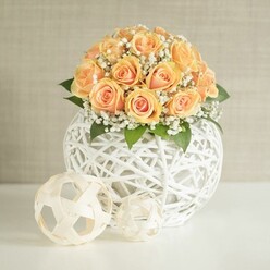 A rounded cluster of peach roses is the star of this image, rising like a gentle dome of warmth from a contemporary white woven vase. Around two dozen rose blooms are packed closely together, each one a study in subtle variation: the inner petals glow with deeper apricot and burnt orange notes, while the outer edges soften to a creamy, almost shell-like peach, giving the bouquet depth and a luminous, candlelit quality. Threaded between the roses are countless sprigs of baby's breath, their tiny white flowers forming a misty halo that lightens the arrangement and introduces a feeling of celebration without ever overpowering it. A few dark green leaves sit low around the base, offering contrast and a reminder of the garden. The vase itself is a striking, spherical piece, formed from white strips woven into an airy lattice, like a modern rattan lantern. Its open pattern casts delicate shadows and allows you to sense the volume of the bouquet rather than hiding it. Resting on the same smooth white surface in front are two smaller decorative spheres, created from similar woven material in soft natural tones, one medium and one petite, mirroring the main vessel in a quieter key. The backdrop is a light, greige wall with fine vertical texture that keeps the focus on the flowers. The arrangement feels like it could be placed on a polished sideboard in a Canonbury terrace overlooking New River Walk, ready to welcome guests for an intimate dinner or to add a refined, floral accent to a freshly styled living room. Crafted with care and a keen eye for texture, it reflects the calm yet sophisticated taste often sought in this corner of North London.