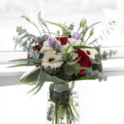 Bathed in gentle daylight much like the soft glow that washes over Canonbury's historic terraces, this bouquet speaks of subtle refinement and heartfelt artistry. The deep red roses, plush and richly coloured, draw the eye and infuse the arrangement with classic romance. Around them, the snowy petals of white gerberas seem almost luminous, each flower's espresso-brown centre lending a touch of grounding warmth. Sprigs of silvery eucalyptus arc gracefully outward, their rounded leaves offering both visual intrigue and a fresh, invigorating backdrop-a familiar scent on walks through Arlington Square in spring. Pale lavender lisianthus, some still in a state of expectant bloom, thread their tender hues among the bold florals, providing a feeling of gentle anticipation. Wisps of fluffy white filler flowers scatter like soft clouds, echoing the tranquil spirit of nearby New River Walks. At the base, glossy, sculpted leaves cradle the bouquet in verdant embrace, their deep green luster highlighting every colour above. The transparent vase allows you a glimpse of water-nourished stems, underscoring the thoughtful freshness Canonbury Flowers brings to the arrangement. With its harmonious blend of texture, colour, and fragrance-a mingling of rose, eucalyptus, and gentle earthiness-this bouquet is as suited to commemorating milestones or sending comfort as it is to brightening a Stoke Newington breakfast nook. Each stem and blossom reflects a local florist's artistry and a deep understanding of the community's understated elegance.