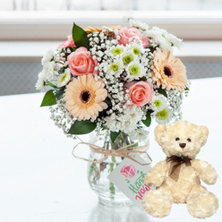On a smooth white surface, a lovingly styled anniversary display captures the feeling of a quiet celebration at home in Canonbury. At the heart of the scene is a compact, rounded bouquet arranged in a clear glass jar vase, its sides catching the light like a small pool of water. The bouquet blends soft pink roses with peach-toned gerbera daisies whose rich dark centres lend depth and contrast. Around them, airy white baby's breath forms a delicate halo, while clusters of small white chrysanthemums with lime-green hearts bring a cool brightness that suggests early spring days in nearby Highbury Fields. A simple band of natural twine encircles the neck of the vase, knotted with an easy elegance. From it dangles a small white tag, printed with a pink graphic and fine green lettering indicating that the gift has been handmade, a quiet reminder of the personal thought behind it. Just to the right, a cream teddy bear sits slightly turned toward the flowers, as if keeping them company. Its fur is downy and inviting, with dark beadlike eyes, a brown nose, and a neatly tied brown satin ribbon giving it a classic, timeless look. Soft daylight floods in from the background, which is intentionally blurred so that only the suggestion of a bright interior remains, perhaps like a sunlit Canonbury kitchen on a weekend morning. The overall mood is one of gentle romance and reassurance, perfectly suited to an anniversary surprise delivered locally, where flowers and a cuddle toy come together to say, "You're still my favourite person."