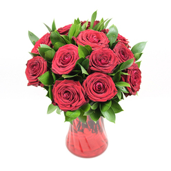 An opulent cluster of deep crimson roses fills the frame, arranged in a translucent red glass vase that seems to glow softly against a pristine white backdrop. There are around a dozen or slightly more roses, each one in perfect bloom, with velvety petals unfurling in tight spirals that reveal intricate layers at the heart of every flower. Their colour is a rich, ruby red, the sort that calls to mind long-held declarations of love and the quiet intensity of an anniversary dinner at home in Canonbury. Between the blooms, a generous amount of dark, glossy greenery - likely ruscus - fans out, the pointed leaves emerging confidently from the cluster of roses. This foliage brings a fresh, woodland contrast, its deep green tones sharpening the drama of the red and giving the bouquet a lush, rounded presence. The vase beneath them curves with graceful simplicity, narrowing towards the base and then opening gently at the rim. Its red glass surface has a subtle ribbed or wavy texture, catching reflections of light that dance up and down the sides like ripples on water. Because the glass is partly transparent, you can glimpse the stems gathered neatly within, adding a sense of honesty and craftsmanship to the composition. With the clean white background and bright, even lighting, the arrangement feels almost sculptural, as though it has been carefully composed by an experienced Canonbury florist who understands how to balance drama with elegance. This image conjures the soft scent of fresh roses and the quiet emotion of a gift delivered across local streets - perhaps arriving at a townhouse off Upper Street or a hidden garden flat near Canonbury Square - to say "I love you" without needing many words.