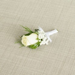 A refined white rose boutonniere is photographed resting on a neutral, linen-like fabric backdrop in a pale beige tone, the woven texture running diagonally beneath the arrangement and giving the scene a natural, understated elegance. At the centre, a single creamy white rosebud is tightly formed yet plush, its outer petals curling inwards as if just about to unfurl, with faint green at the base where the sepals clasp the stem. Surrounding this central bloom, clouds of delicate white baby's breath create a soft, misty frame, their many tiny blossoms forming a lacework of texture that feels light and romantic. Slender, glossy green leaves curve gently around the rose, joined by a few finer, fern-like sprigs that add movement and depth, suggesting the careful hand of an experienced florist. The stems are smoothly wrapped in a bright white satin ribbon, its surface catching a subtle sheen in the soft, diffused light. A small, precise bow sits just under the flower heads, its loops neat and balanced, making it clear that this is designed to sit smartly on a suit or tuxedo lapel. The overall composition is compact and tailored, ideal for a groom walking from a Canonbury townhouse toward an intimate ceremony in a local church or a hidden garden near the New River Walk. The atmosphere of the image is quiet and thoughtful, as if captured in the calm moments before guests arrive, with the boutonniere symbolising purity, new beginnings, and the gentle formality that suits weddings and black-tie events across this tranquil North London neighbourhood.