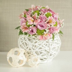 Envisioned for a sophisticated setting in the heart of Canonbury, this floral arrangement merges contemporary artistry with heartfelt, handcrafted warmth. The centerpiece blossoms with pastel pink and lavender roses, each petal lush and velvety, their colors softly shimmering in the daylight that pours through North London windows. Pink alstroemeria-Peruvian lilies-add a gentle, stripy intrigue with fluted petals marked by fine hints of yellow and deep pink. Lime green button chrysanthemums-cheerful and playful-cluster among the blooms, introducing a pop of vivid freshness. Wisps of white baby's breath lace the bouquet, imparting a lightness that dances in the gentle air. Deep, glossy green leaves peek from beneath, adding natural depth and anchoring the palette. The entire bouquet is cradled within a remarkable white vase whose intricate, open-weave lattice hints at artisanal craftsmanship borrowed from Canonbury's thriving creative scene. Its generous, rounded shape offers sturdy support, while a pair of decorative spheres-woven from pale, natural strands-are thoughtfully placed to the left, echoing the vase's texture and adding a harmonious flourish to the composition. The image's soft beige background and luminous white table let the bouquet's colours and sculptural shapes shine, conjuring an inviting, calming atmosphere perfect for a wedding table, a cherished anniversary, or as a statement piece for homes along Canonbury's leafy avenues. The entire scene exudes assurance and gentle joy, making this arrangement a memorable choice for celebrating life's meaningful moments locally.
