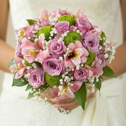 Held with tender grace and local charm, this Canonbury bridal bouquet reveals a masterful interplay of colour, texture, and emotion. The main focus falls on softly lilac roses and gentle pink blossoms, their hues radiating comfort and understated sophistication-just right for a wedding in one of Canonbury's leafy garden squares. Bright green button chrysanthemums nestle between, their tidy orbs lively and crisp against the velvety spiral of the rose petals, while delicate alstroemeria introduce gentle streaks of maroon and golden yellow, bringing depth and subtle fragrance. Baby's breath is woven artfully throughout, its fine white clouds providing softness that gently ties together the bolder tones. Supporting leaves-deep, glossy green-peek out at the edges, grounding the design with vitality reminiscent of spring afternoons strolling along the New River Walk. As a Canonbury florist, I see in this bouquet a true harmony between tradition and fresh beginnings, perfect for a radiant bride and a celebration tinged with heartfelt promise. Each element speaks to craftsmanship and a sense of place, ideal for couples seeking wedding flowers that reflect the unique charm of North London.
