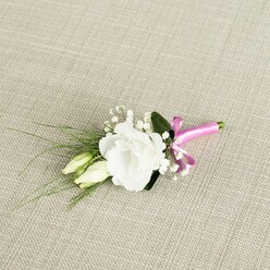 A refined white lisianthus boutonniere rests at a gentle diagonal on warm, light beige fabric, the weave reminiscent of natural linen you might find in a Canonbury artisan homeware shop. The fully open lisianthus flower is the clear focal point, its soft white petals layered in delicate ruffles that curve outward from a calm, luminous centre. Just beneath and to the side, two slender lisianthus buds in a creamy, pale green hue lean protectively against the main bloom, their smooth surfaces and closed tips hinting at blossoms yet to come. Sprinkled among them are airy stems of white baby's breath, each tiny flower no bigger than a pinhead, catching the light like small stars and adding a lace-like softness to the composition. From behind the central bloom, fine, grass-like leaves sweep outwards towards the lower left corner, their deep green tones and slender lines offering movement and a sense of natural growth. A broader, darker green leaf is tucked discreetly at the back, anchoring the arrangement and adding a quiet richness to the palette. At the stem ends, a bright fuchsia satin ribbon wraps everything neatly together, tied into a small bow that gleams against the matte texture of the fabric, introducing a joyful, contemporary pop of colour to the otherwise classic white and green scheme. The soft, even lighting reveals each petal edge and leaf vein without harsh shadows, giving the impression of a carefully handcrafted piece, ready to be worn at a Canonbury wedding, an intimate ceremony off Upper Street, or an elegant formal event, carrying the gentle freshness of early spring.