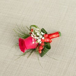 On a pale beige, finely woven fabric, a single statement boutonniere is displayed with the care of a still-life painting, its colours echoing the quiet sophistication of weddings held around Canonbury's leafy squares. The focal flower is a lush fuchsia rose, its petals dense and velvety, cupped tightly at the centre and then opening outward in layered swirls that suggest both tenderness and confidence. Surrounding the rose are airy tufts of white baby's breath, dozens of miniature blossoms clustered like soft foam, bringing a sense of lightness and purity to the composition. Tucked among these are small, polished red berries, reminiscent of hypericum, which glow like tiny lanterns against the greenery and intensify the warm, celebratory mood. A combination of slender, fern-like greenery and more substantial dark green leaves fan out from the base, each leaf carefully curled and positioned to create depth and a graceful, tapered silhouette that would sit neatly on a lapel. The stems are expertly wrapped in a rich red satin ribbon, the fabric smooth and gleaming as it coils tightly around the base and finishes in a delicate, symmetrical bow. Soft natural light, as if from a nearby Canonbury window on a quiet afternoon, bathes the arrangement, enhancing the textures of petal, leaf, and cloth. This thoughtfully composed floral accessory feels perfect for a groom, best man, or prom guest, its intimate scale and harmonious palette expressing joy, romance, and the kind of refined detail that a local florist brings to special occasion flowers in North London.